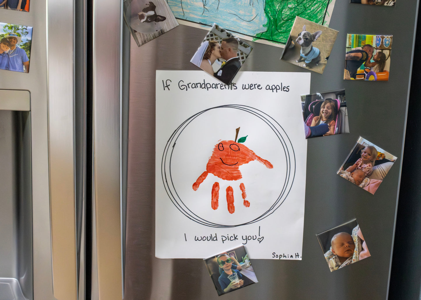 Handmade card on a refrigerator with handprints and text, surrounded by family photos.