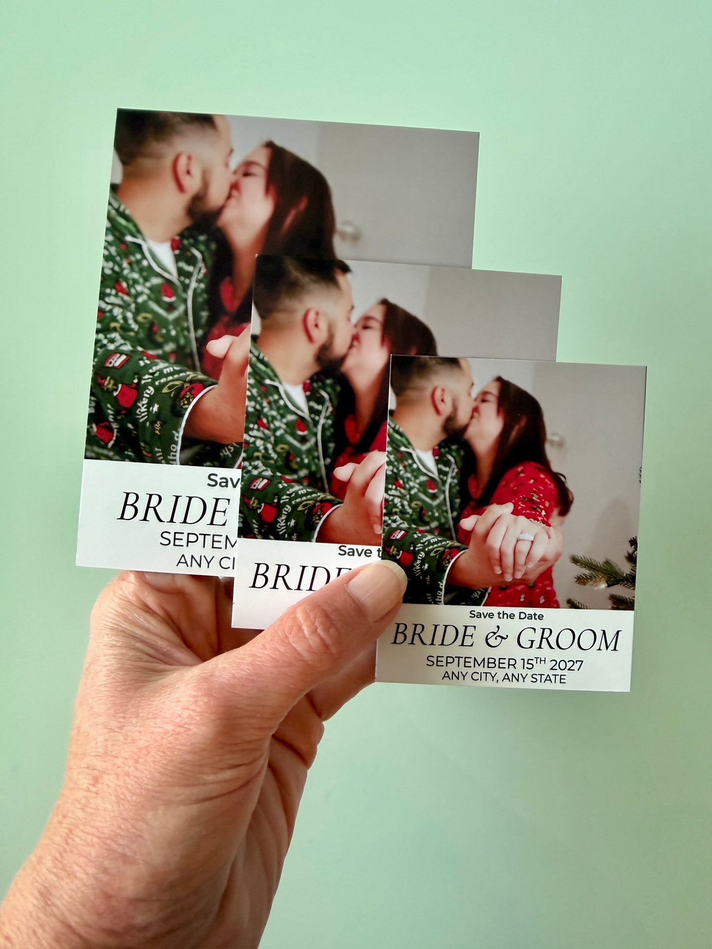 Hand holding a set of 'Save the Date' cards with a couple in Christmas-themed outfits against a light green background.